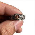 Close-up of a hand-forged copper ring with a rough rock texture and a detailed Buddha motif, shown on a finger.
